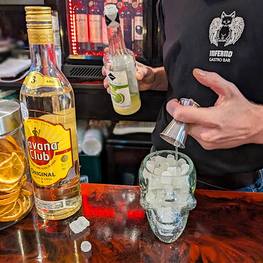 Bartender from Inferno Gastro Bar preparing a daiquiri, adding Havana Club rum and lime juice in a skull-shaped glass filled with ice.
